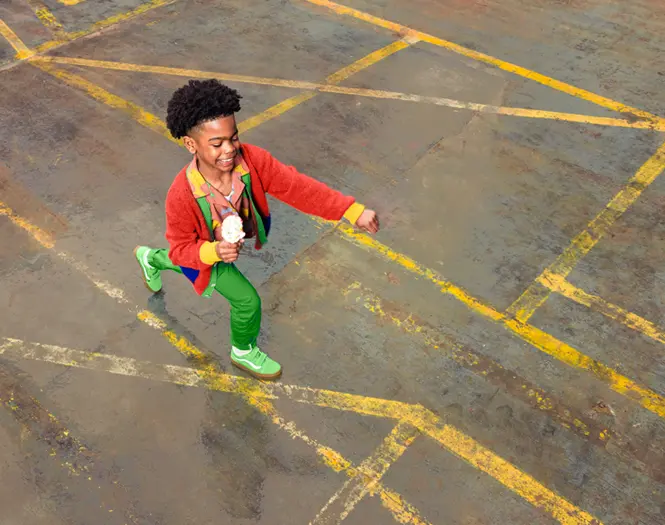 a young boy wearing a red jecket and green pants walking with an ice cream cone in his hand